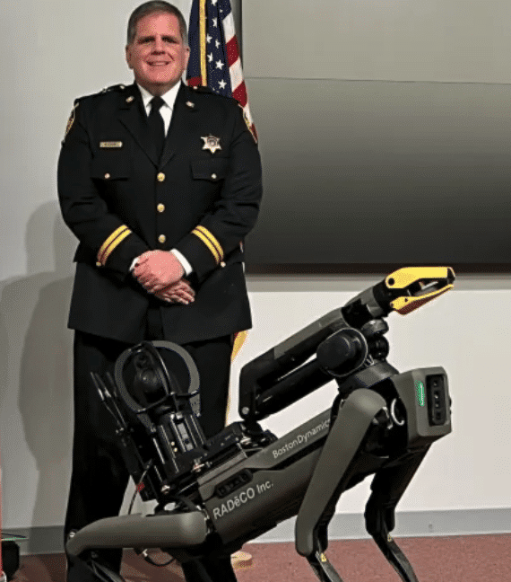 A gray Spot robot with arm and thermal camera poses with a uniformed police lieutenant